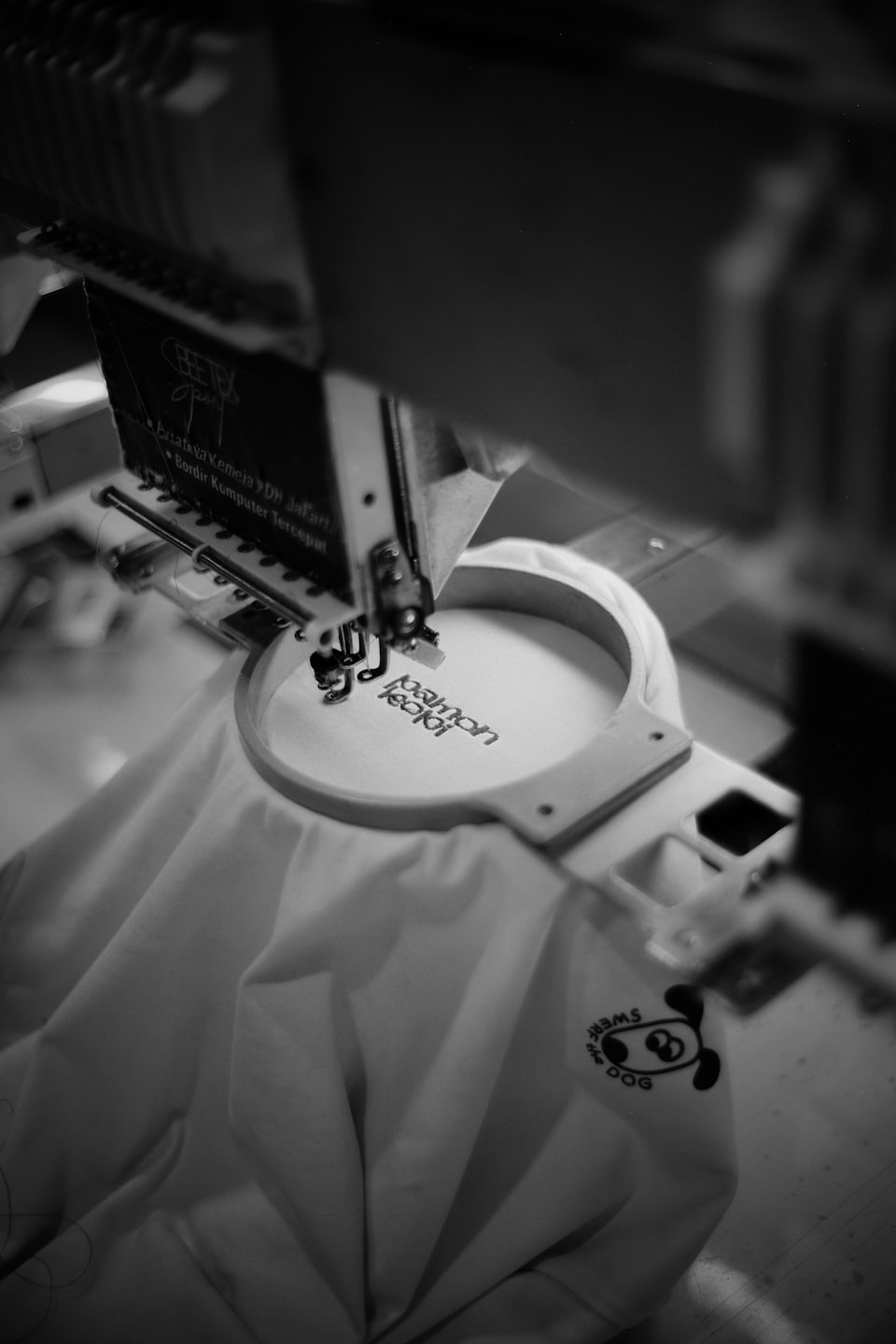 Una máquina de bordar industrial cose texto sobre tela tensada.